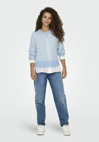 Maglione lavorato a maglia blu chiaro con motivo a onde, indossato sopra una camicia bianca, abbinato a jeans blu larghi e sneakers bianche.