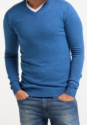 Pull en tricot bleu avec un col en V, à manches longues et des poignets côtelés. Porté par-dessus un t-shirt blanc, associé à un jean en denim.