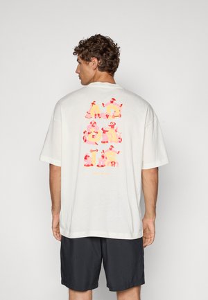 Camiseta de color crema claro de corte oversize con un gráfico colorido en la parte posterior, con un ajuste relajado y mangas cortas. Combinada con pantalones cortos oscuros.