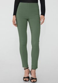 Olivově zelené legíny s vysokým pasem vyrobené z elastického materiálu, s elegantním designem a přiléhavým střihem, doplněné černými sandály na řemínky.