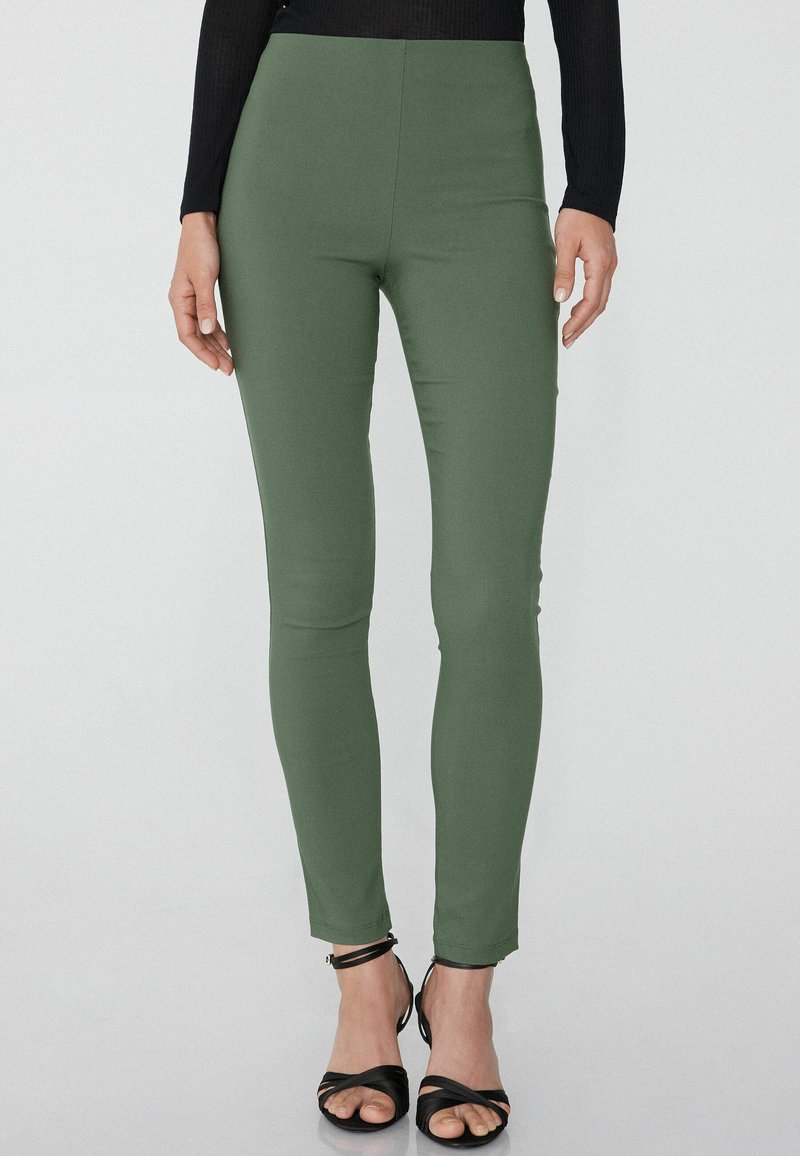 Olivově zelené legíny s vysokým pasem vyrobené z elastického materiálu, s elegantním designem a přiléhavým střihem, doplněné černými sandály na řemínky.