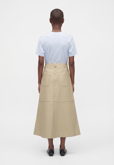 Personne portant une chemise bleu clair à manches courtes et une jupe midi beige taille haute avec des poches arrière, debout de dos.