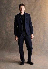Marint blazer med en ficka, svart polotröja, mörka denimjeans och svarta läderskor. Slätt tyg och skräddarsydd passform.