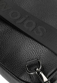 Czarna skórzana torebka crossbody z teksturowaną powierzchnią, plecionym paskiem z wytłoczonym logo oraz metalowym zapięciem.