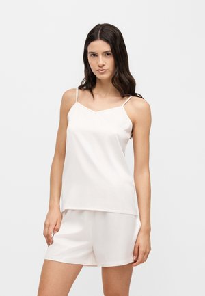 Vrouw met lang donker haar, gekleed in een lichtkleurige mouwloze top en bijpassende korte broek tegen een effen witte achtergrond.