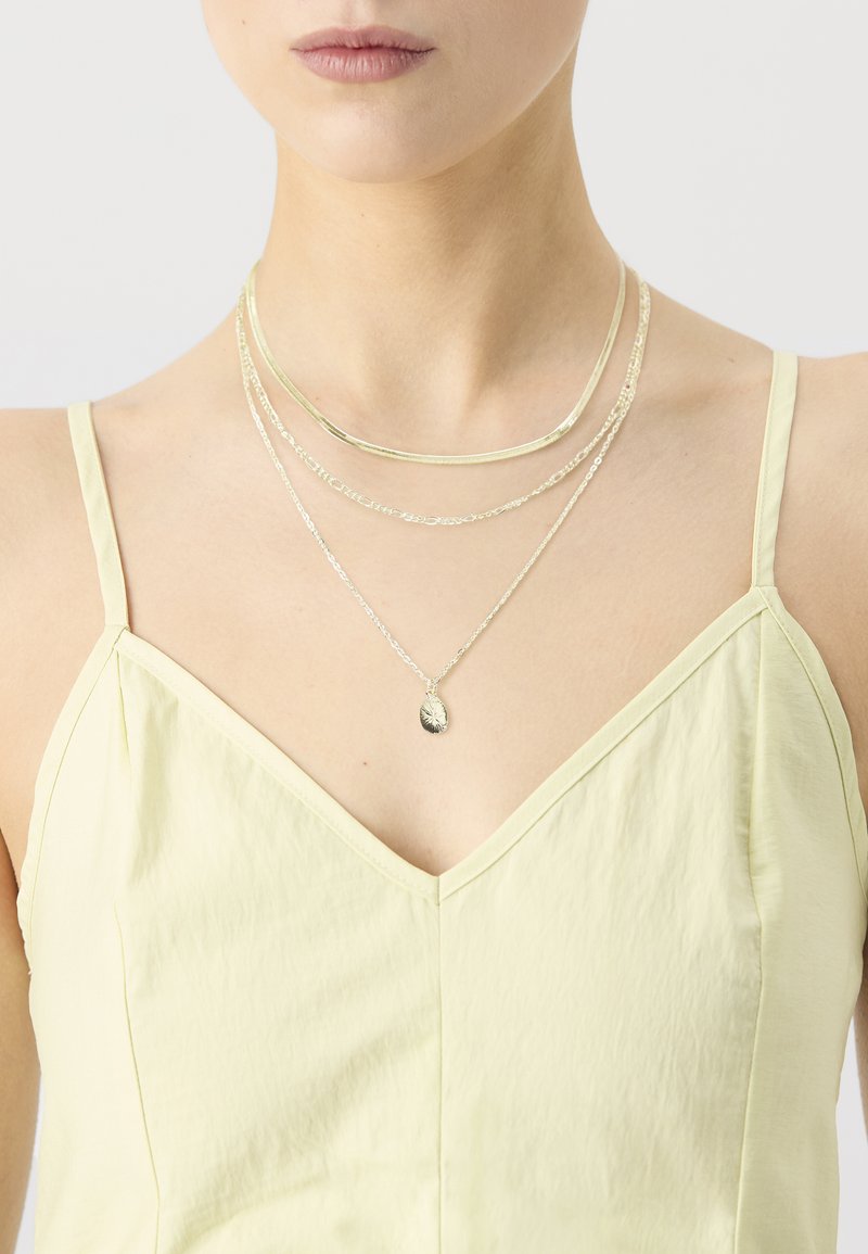 Des colliers superposés en argent avec une fine chaîne, des surfaces métalliques lisses et un pendentif ovale, portés par un modèle avec un haut jaune clair.