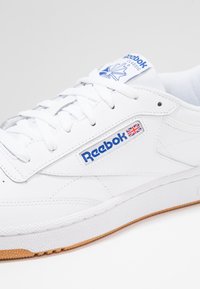 Adidași Reebok albi cu un superior din piele netedă, detalii cu logo albastru și talpă din cauciuc de culoare caramel. Dispune de șireturi albe și guler căptușit.