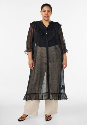 Kvinde iført en gennemsigtig sort knap-kjole med flæser over beige bukser med vide ben og sorte hælede sandaler, stående mod en hvid baggrund.