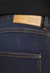 Tamsiai mėlynos džinsinės kelnės su oranžiniais siūlais, ant liemens juostos yra rudas odinis etiketė, kurioje rašoma "DENIM KELIONĖ."
