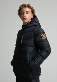 Έφηβος με σγουρά μαλλιά, φοράει μαύρο μπουφάν με κουκούλα και το λογότυπο ZAVETTI CANADA στο μανίκι, κοιτάζει την κάμερα.