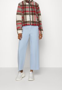 Giacca a quadri accorciata nei toni del marrone, rosso e crema, con colletto alto e zip frontale. Pantaloni a gamba larga blu chiaro e sneakers bianche.