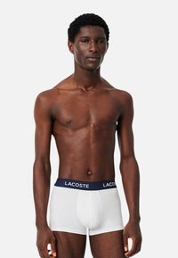 Fehér boxer alsónadrág sötétkék elasztikus derékpánttal, amelyen a "LACOSTE" logó található. Simaság jellemzi, testhezálló kialakítás, látható varratok nélkül.