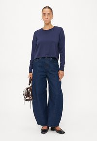 Felpa corta blu navy abbinata a jeans svasati in denim scuro, con una borsa marrone e scarpe basse nere. Elementi di design semplici.