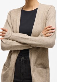 Beige knitted cardigan with open front, two front pockets, ribbed cuffs, and a soft texture, paired with a black fitted top and dark pants.