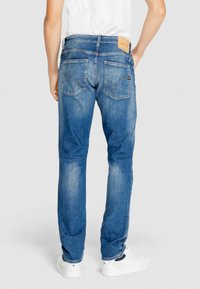 Jeans de mezclilla azul con corte recto, con bolsillos traseros y una etiqueta de cuero en la cinturilla. Usados con zapatillas blancas.
