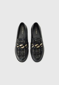 Musta loafers, joissa on tekstiilistä valmistettu päällinen, joka sisältää kultaketjuakun instepin yli, sileät nahkayksityiskohdat ja litteät pohjat.