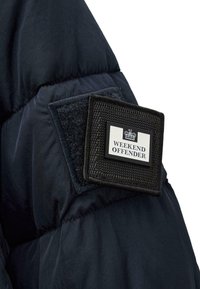Marineblå pufferjakke-sektion med et sort stofmærke med "WEEKEND OFFENDER" i hvid tekst og et krone-logo, fastgjort med velcro.