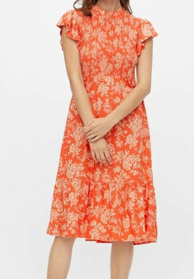 Femme portant une robe orange mi-longue avec des motifs floraux crème et des manches courtes volantées, debout les mains croisées à la taille.