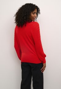 Pull rouge avec des manches texturées et côtelées et un corps lisse. Présente un col rond et des manches retroussées. Porté avec un pantalon sombre.