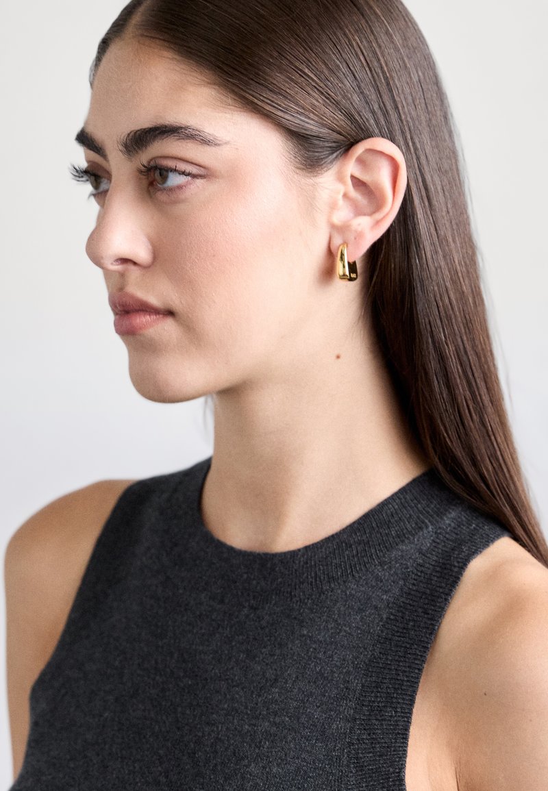 Jauna sieviete ar taisniem brūniem matiem, tērpusies tumšā bezpiedurkņu topā, ar mazām spīdīgām zelta auskariem gredzenos, skatās pa kreisi uz vienkārša fona.