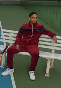 Sarkans velveta sporta uzvalks ar zilo svītru, baltas kedas, sēž uz baltām soliem zaļā tenisa kortā. Tenisa rakete blakus.