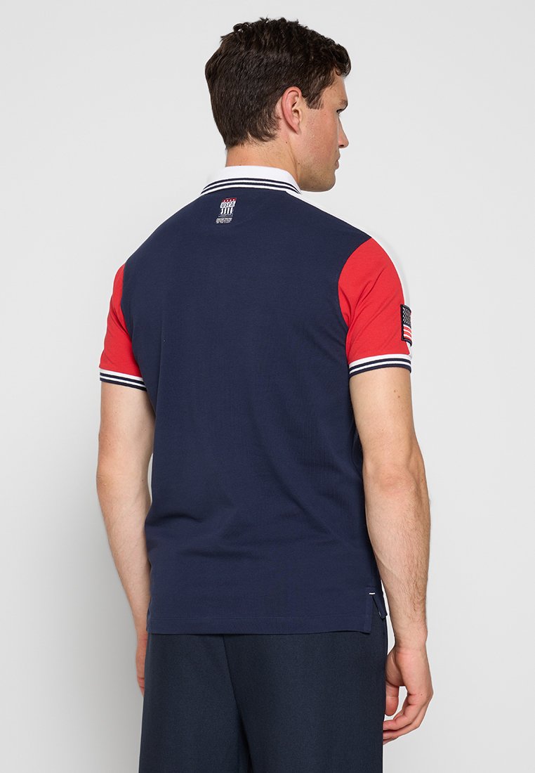 T-shirt polo avec un corps bleu marine, des manches courtes rouges, un col rayé et un écusson représentant un drapeau américain sur la manche. Texture lisse et côtelée.