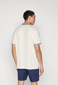 Κρεμ πόλο μπλούζα με κοντά μανίκια, γκρι γιακά και λεπτομέρειες στα μανίκια· διαθέτει ίσιο τελείωμα και πλαϊνές σχισμές για ευκολία στις κινήσεις.