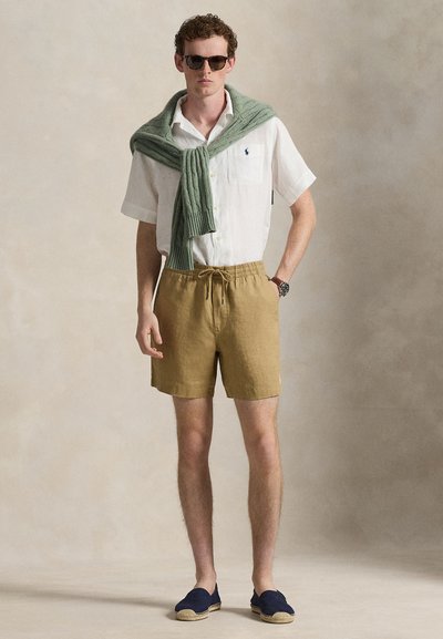 Λευκό πουκάμισο με κοντά μανίκια, χακί σορτς, πράσινο πλεκτό πουλόβερ δεμένο στον λαιμό, καφέ εσπαντρίγιες και στρογγυλά γυαλιά ηλίου. Απλό φόντο.