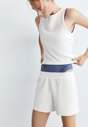 Nő fehér ujjatlan felsőt és fehér, rugalmas derékpántos rövidnadrágot visel, és igazítja a rövidnadrágját, hogy láthatóvá váljon a kék fehérnemű derékpánt.