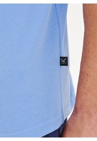 Jasnoniebieska koszulka z bawełny, z czarną metką z krzyżowym wzorem na dolnej stronie oraz gładką, miękką teksturą.