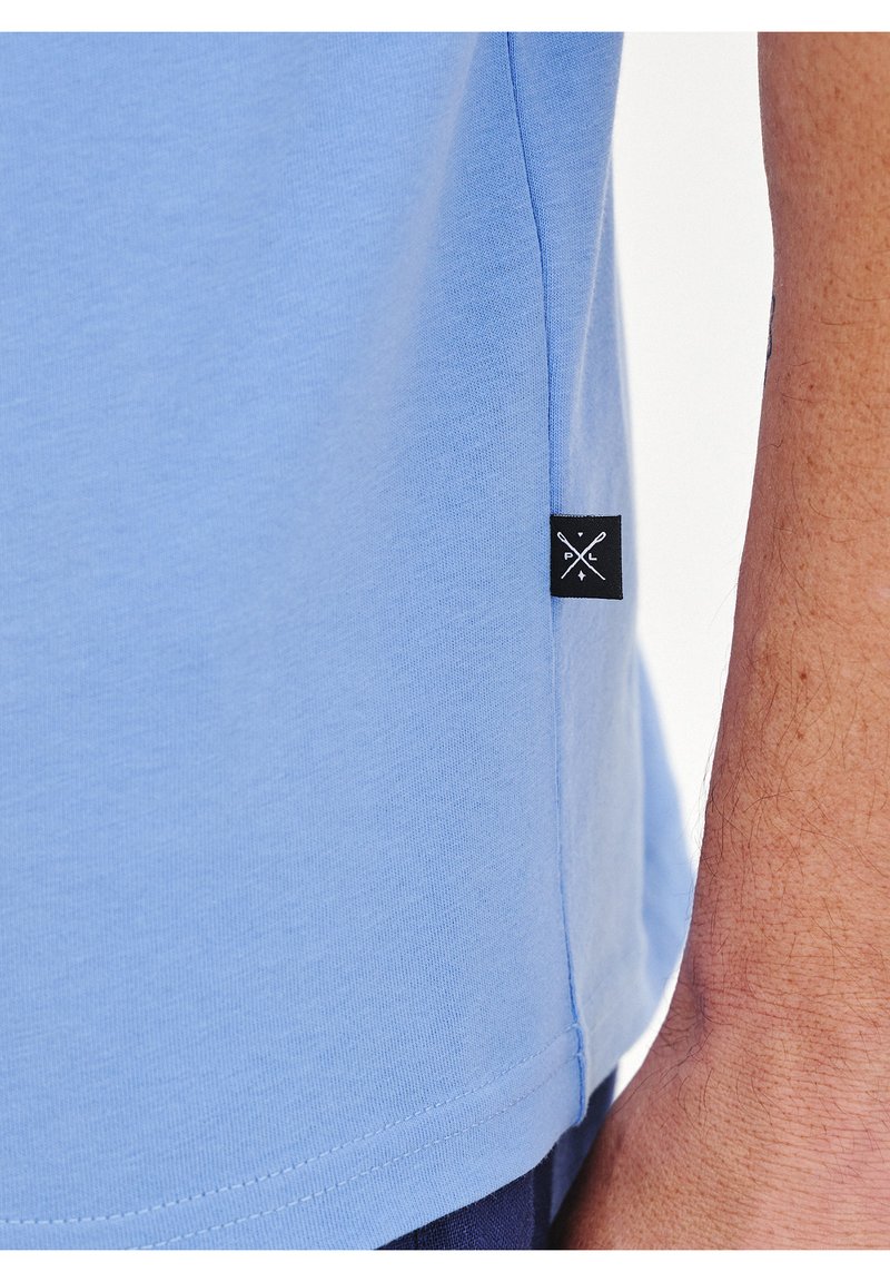 Jasnoniebieska koszulka z bawełny, z czarną metką z krzyżowym wzorem na dolnej stronie oraz gładką, miękką teksturą.