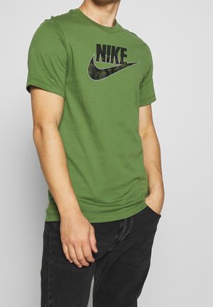 Mężczyzna w zielonej koszulce Nike z krótkim rękawem z czarnym logo i czarnych spodniach, stoi z jedną ręką w kieszeni na jednolitym tle.