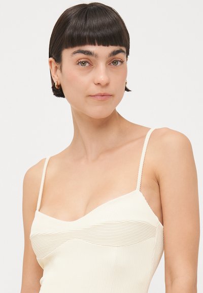 Helles beige Tanktop mit gerippter Textur, dünnen Trägern und einem geschwungenen Ausschnitt. Verfügt über horizontale Nahtakzente über der Brust.
