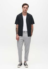 Schwarze kurzärmelige Hemdbluse, weißes T-Shirt darunter, hellgraue schmal geschnittene Hose und schwarze Sneakers mit weißen Sohlen, vor einem weißen Hintergrund stehend.