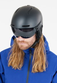 Ski-hjelm i mat sort med indbygget visir, der har ventilationshuller og justerbare stropper. Kombineret med en blå teknisk jakke.