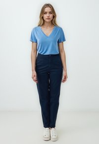 T-shirt blu a scollo a V, abbinato a pantaloni blu scuro e sneakers bianchi. Tessuto morbido, design aderente e dettagli con bottoni visibili sui pantaloni.