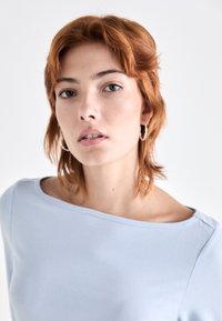Lys blå langermet topp med bred utringning, laget av glatt stoff. Modellen har kort, uregjerlig rødlig hår og små hoops-øreringer.