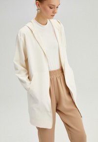 Jachetă cu fermoar de culoare crem deschis, cu croială lejeră, având două buzunare frontale și glugă cu șnur, asortată cu pantaloni bej cu talie elastică.