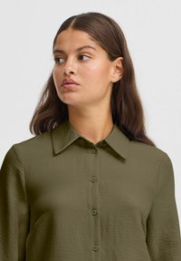 Olive green button-up shirt with a textured fabric, featuring a collar and a front placket with buttons, tailored fit.
