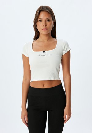 Crop top blanc à côtes avec un décolleté carré, des manches courtes et un détail de nœud noir au centre, présentant un texte brodé en dessous.