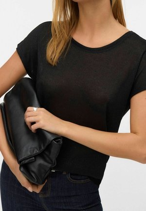 Vrouw met een zwart kort mouwtopje en blauwe spijkerbroek houdt een grote gevouwen zwarte leren clutch vast tegen een witte achtergrond.