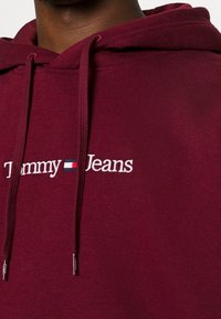 Felpa con cappuccio color bordeaux realizzata in tessuto morbido, caratterizzata da un logo ricamato "Tommy Jeans" in bianco e blu, con lacci sul cappuccio.