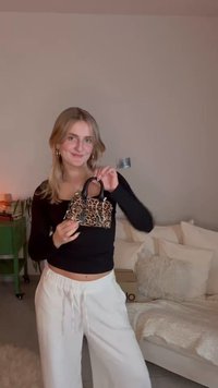 Leopardikuvioinen minikäsilaukku, jossa mustat kahvat ja hopeiset metalliosat. Laukulla on jämäkkä muoto ja kompakti design, ja sen pitelee malli.