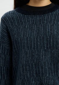 Tumši zili tekstilizēta adīta džemperis ar vertikālu ribotu rakstu, valkāts virs melna apaļā kakla krekla, saīsināts zem zoda.