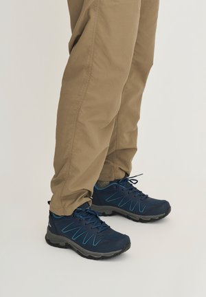 Beine, die beige Hosen und dunkelblaue Wanderschuhe mit schwarzen Sohlen tragen, vor einem einfarbigen hellen Hintergrund gezeigt.