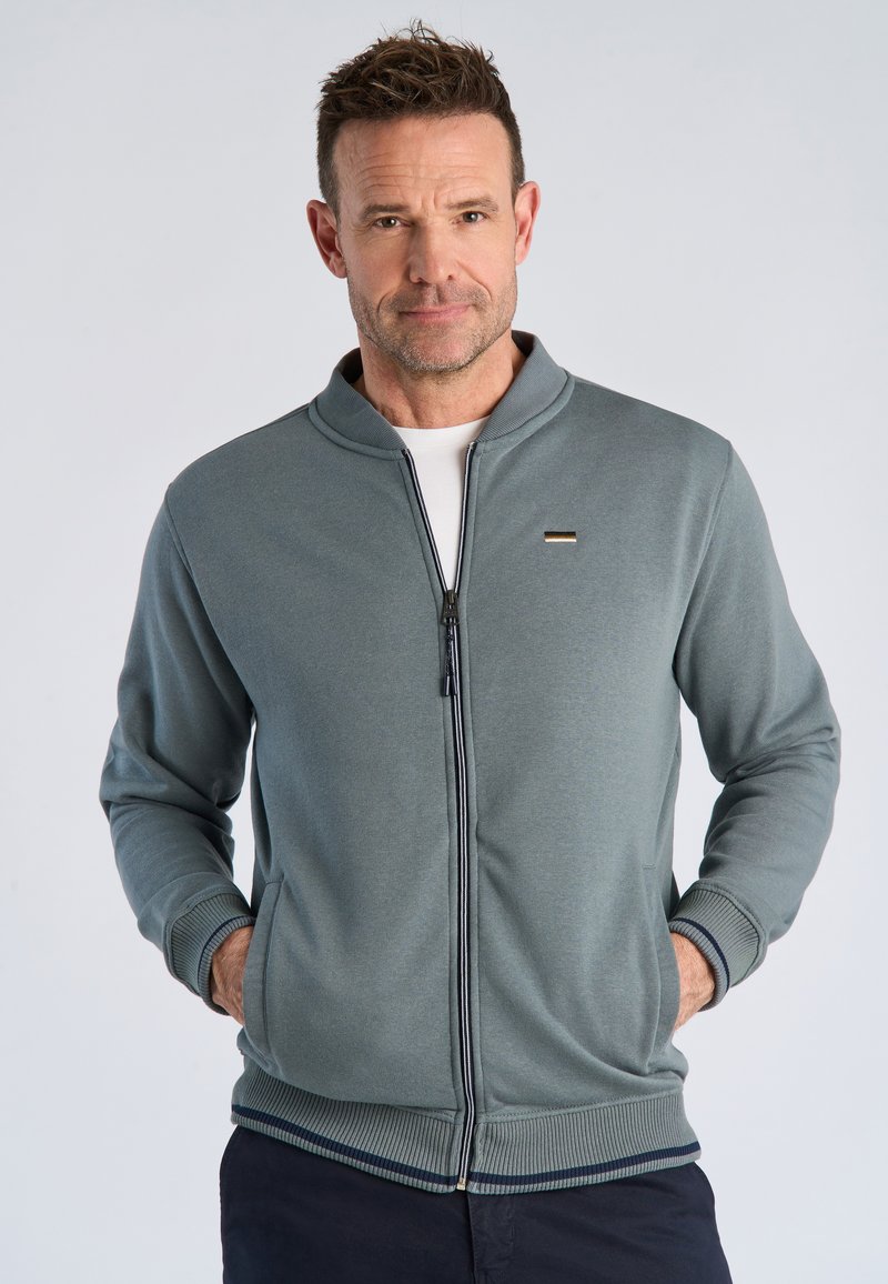 Mand iført en grå lynlåstrøje over en hvid skjorte med hænderne i lommerne, stående mod en ensartet lys baggrund.