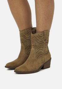 Botas de tobillo de ante en color beige con punta afilada, diseño decorativo perforado, costuras curvadas y tacón apilado. Cierre con cremallera en el lateral.