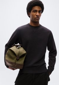Άνδρας με afro κόμμωση, φορώντας μαύρο φούτερ, κρατώντας μια τσάντα duffel σε ελιά πράσινο και καφέ χρώμα, μπροστά σε απλό φόντο.