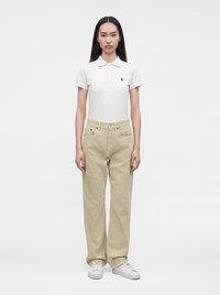 Femme aux longs cheveux noirs debout face à l'avant, portant un polo blanc à manches courtes, un pantalon beige et des baskets blanches sur un fond uni.