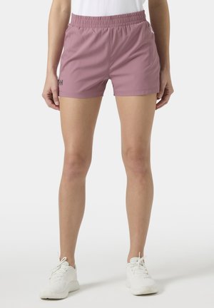 Mujer con pantalones cortos deportivos elásticos de cintura color malva y zapatillas blancas, de pie contra un fondo claro y liso.